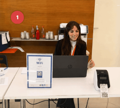 Pessoa em uma mesa de credenciamento com laptop, impressora e QR Code para registrar participantes.